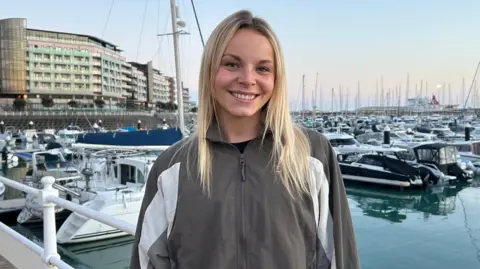 Holly Le Feuvre stands outdoors in front of the marina filled with numerous docked boats and masts. She is wearing a grey and white zip-up jacket, has blonde hair, and is positioned near a white railing. Behind the marina, a large modern building stretches along the waterfront.