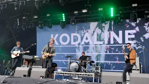 Kodaline: Irish rock band to split after over a decade 4 Kodaline play on stage. There is a banner behind them which says 'Kodaline'.