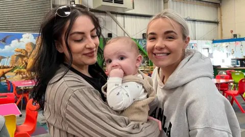 Two women are smiling. One of them, who has long, dark hair and is wearing a beige top, is holding a baby. The second woman has blonde hair and is wearing a grey hooded sweatshirt.