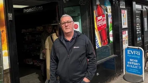 Kevin Shoesmith/BBC A man in his mid 60s, with grey short hair and glasses, is standing in front of a convenience store with a black frontage and murals showing brands such as Coca-Cola, Weetabix and Fairy. A sign for Evri parcelshop stands on the pavement.