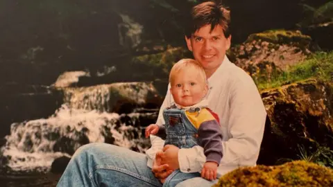 Family photo An archive picture of Dr Nicholas Bennett when his son Alexander was a young child. They are sitting by a waterfall. The father is holding the child in his lap.