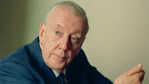 Fritz Curzon Malcolm Arnold, looking to the left, with short grey hair, wearing a blue tie, white shirt and