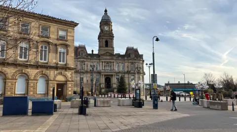 Image shows Dewsbury town centre. The town hall is in the background, a big stone building.
To the left of shot is also a boarded up , stone building which used to be Barclays bank but has stood empty for the last two years. 