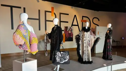 In this photograph there are four Mannequins. The first Mannequin is wearing a thick multi-coloured knit cardigan. The second Mannequin is facing a different direction wearing a black gown with a slim black scarf on and hanging at the bottom of their arm is a large tote bag. The third mannequin is wearing a light-coloured gown with a dramatic frilled black colour around the neck and hems. The fourth one is wearing a long black floral gown.