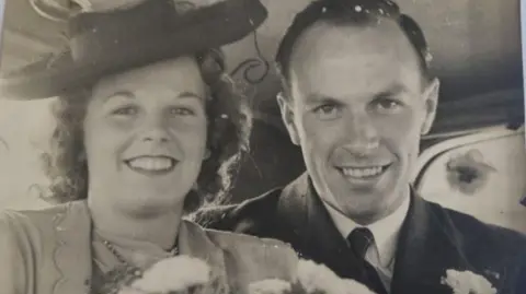 Marjorie O'Hanlon Man and woman are sitting in a car and both are smiling. Man wears a suit with tie. His wife has a hat, carries flowers and is also in a light-coloured suit. The image is a black and white photo.