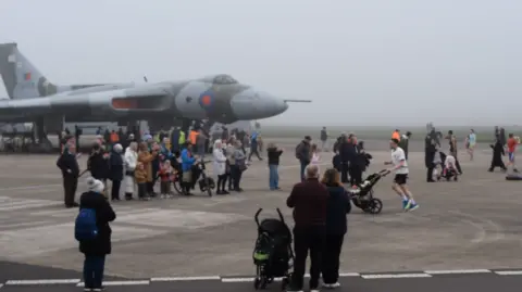 Yasper A runner pushing a pram next to a fighter plane. A small crowd of people is gathered to applaud his efforts.