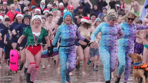 Medios de comunicación de PA: Tres mujeres corren hacia el mar vestidas de pez con pijamas de escamas azules y moradas. Se ríen y hay un labrador dorado con chaleco salvavidas a su lado. Hay una multitud sonriendo y corriendo detrás de ellas, incluyendo a una mujer rubia vestida de elfo a su izquierda.