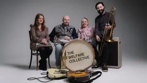 Four members of The Welfare Collective's horn section. From left, Philippa Smith is sitting and holding a cornet in her lap, James Wright is sitting and has a euphonium, Jo Wright is sitting with a flugel while David Smith is perched on a speaker holding a trombone. A drum and two guitars are placed in front of them. The photograph has been taken against a plain backdrop.