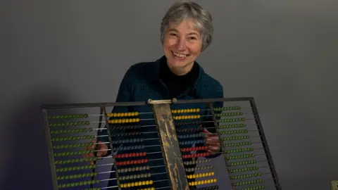 Gillian Cook stands behind a large abacus approximately 80 cm wide. It has several rows of beads of different colors that were used by examiners to determine grade standards.