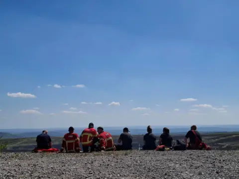 West Yorkshire Fire and Rescue Service Eight people are picture seated on the ground overlooking moorland. Three of them are wearing red jackets with yellow, fluorescent stripes and the word 'Fire' on the back.