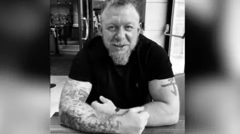 A black and white photograph of a man sitting at a table and smiling. He appears to be in a restaurant or bar. He has his arms resting on the table in front of him and has lots of tattoos on one arm and one tattoo on the other. He has a medium-length beard and short hair and is wearing a black, fitted T-shirt.