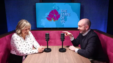 Declan Harvey points towards Tara Mills who are both sitting in a podcast Studio. There is a television screen with the special logo for the state of us podcast visible on a wall in the background. They are seated on a pink sofa. Tara is wearing a white blouse and Declan has a black jumper on.