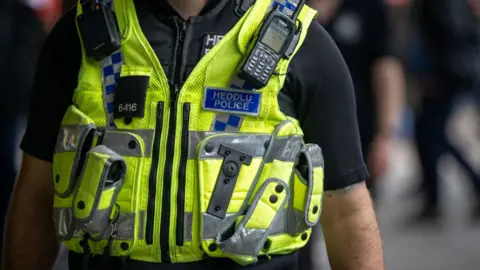 A police officer on August 12, 2023 in Cardiff, Wales. He is wearing a luminous yellow vest with Heddlu written on the back in white on a blue background.