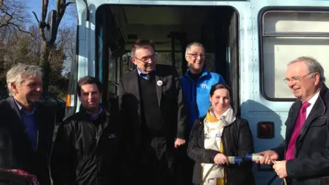 BBC Alex Townsend and invited guests standing in front of the bus while a cable is unplugged