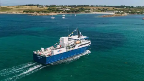 A large blue vessel heads towards the Isles of Scilly across blue waters with boats and land seen in the background 