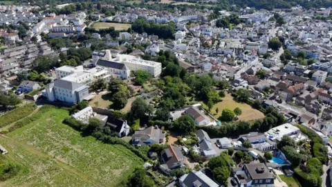 An aerial view of a suburban area with numerous houses, buildings, and greenery. The image shows a mix of residential homes, larger buildings, and open green spaces. The landscape is hilly with clusters of trees and vegetation interspersed among the structures.