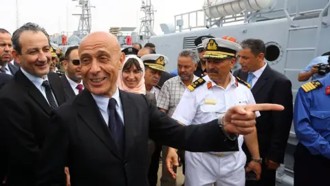 Getty Images Marco Minniti laughs alongside Libyan Defence Minister (of the UN-backed government) at the harbour next to some Libyan coast guard vessels, while naval personnel can also be seen nearby