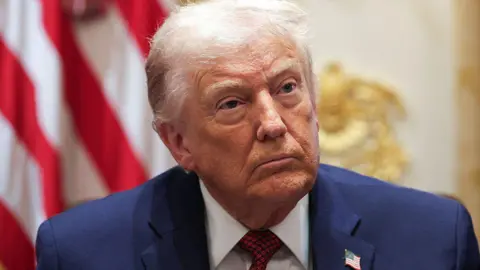 Getty Images Donald Trump sits indoors wearing a dark blue suit and striped red tie, looking slightly upward with a serious expression. A blurred US flag is visible behind him.