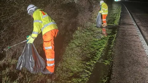 National Highways Two people in hi-vis clothing, helmets and gloves and holding large black rubbish bags and sticks, on a verge by the side of a road.