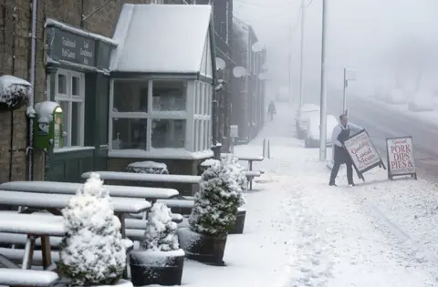 PA Media A local butcher carries his shop sign across a snowy pavement in Tow Law, County Durham, as Storm Eunice sweeps across the UK on 18 February 2022