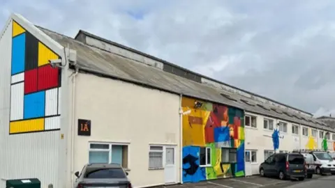 Lansdown Art Studios Association A long warehouse type building with colourful murals on the walls and some cars parked at the front