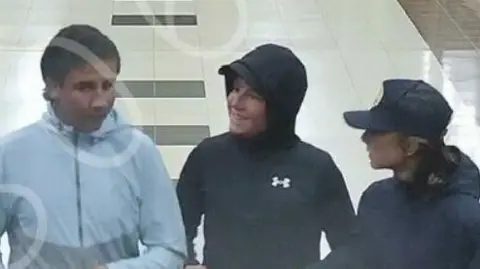 A grainy CCTV still of three boys walking through a shopping centre. They are wearing hooded coats and talking to each other.