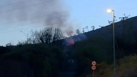 Divis mountain with fire blazing and smoke rising. There is a sign that says 30 (miles per hour). 
