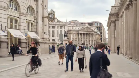 Simon Kennedy People walking in the middle of the road along Threadneedle Street. A cyclist also passes by.