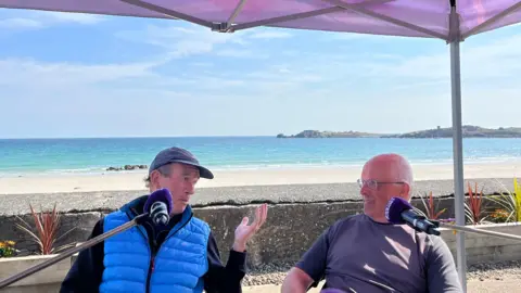 Two men in front of microphones discussing politics, the man on the left is wearing a blue gillet and is gesticulating towards the man on the right. They are in front of a beach backdrop on a sunny day. 