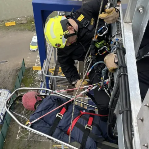 Humberside Fire and Rescue Service Sam hanging out of the window of a training tower while wearing a yellow hi-visibility hard hat and safety gear. He is lowering a dummy body in a metal harness. 