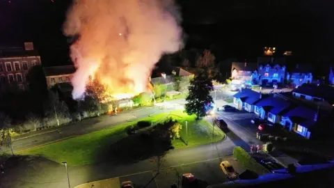 Steven Hooks An aerial photo of Sinton's Mill in Tandragee on fire on Tuesday night. A large plume of smoke and bright orange flames are rising from the Victorian brick complex which is in a housing estate. Firefighters hoses are stretched across the green in front of the mill. Houses and cars are in the foreground.