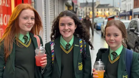 Three teenage girls standing beside each other. The girl on the left has long red hair, the girl in the middle has long curly black hair and the girl on the right has brown hair, tied up. They are all wearing green school uniforms. The girl on the left and the girl on the right are holding drinks in clear plastic cups.