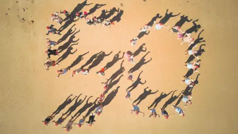 Jersey Long Distance Swimming Club A drone shot from above of several dozen people in bathing suits and red swimming caps standing on a sandy beach spelling out the digits 50.