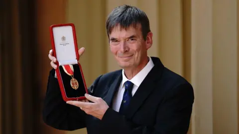 Getty Images Sir Ian Rankin in dark suit and white shirt with blue tie holding up his knighthood. He is smiling. He has dark, straight hair.