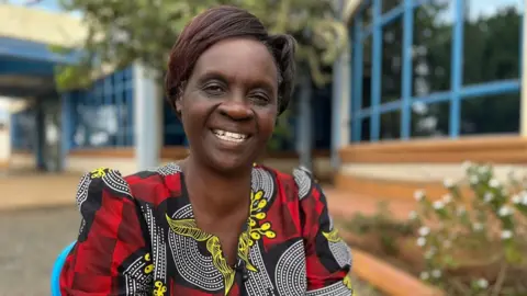BBC A woman in a red, black and white shirt smiles at the camera