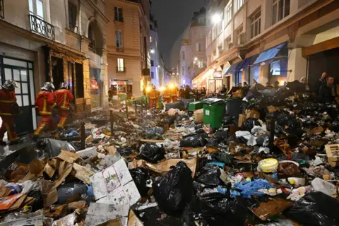 STEFANO RELLANDINI/Getty Images Firefighters checks rubbish after extinguishing a fire during a demonstration, a week after the government pushed a pensions reform through parliament without a vote, using the article 49.3 of the constitution, in Paris on March 23, 2023. - Some 1.089 million protesters took part in demonstrations in France on March 23, 2023, against President Emmanuel Macron's pension reform, the interior ministry said, with 119,000 marching in Paris alone