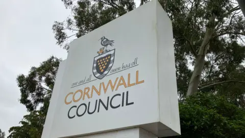 BBC A large white sign with the words 'Cornwall Council'. There is a badge above the text. Behind the sign are trees.