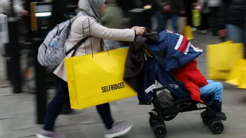 A woman wearing a hijab pushes a pram while carrying a large yellow Selfridges bag down the street.