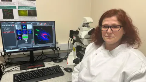 Dr Kirsten Riches-Suman in a lab coat in front of a computer and microscope.