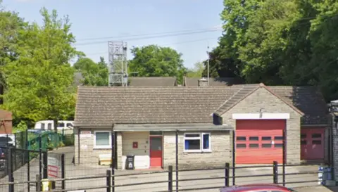 The exterior of Whaley Bridge fire station - a single storey building with two red shutters, a red front door and two white framed windows. The building is surrounded by a black metal fence.