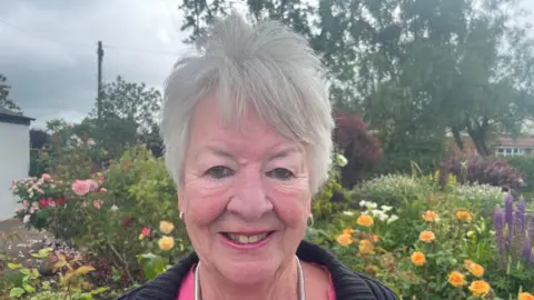 An image of Margaret Smith who is smiling. She has grey hair and is standing in front of a rose bush. She is wearing a pink top and a black cardigan. 