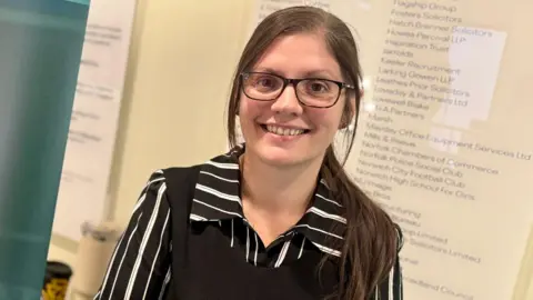 Supplied Charlotte Notley smiles at the camera. She is wearing a black and white striped shirt with a black jumper on top. She has dark glasses and is standing in front of a board with names on it. 
