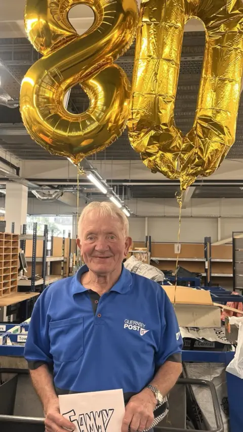 A man with white hair wearing a blue polo shirt, holding a birthday card and stood in front of 2 gold balloons that spell out '80'