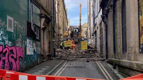 A cordoned off road with three men in hi-vis jackets working on a pile of rubble with a yellow excavator behind them 