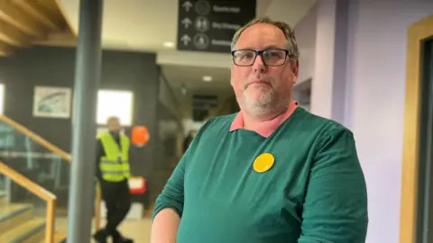 Aisha Iqbal/BBC A man in a green jumper, wearing a yellow Lib Dem badge, stands with arms folded in what looks like a community hall foyers. A security guard stands in the background. 
