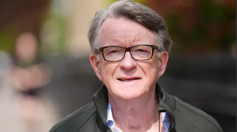 Mandelson pictured walking down a London street. He is dressed casually in a green raincoat and an open necked shirt. 