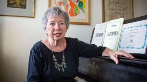 Getty Images An older woman with short greying hair sits at a piano in her home. She is wearing a dark blue top with a silver and turquoise necklace. 