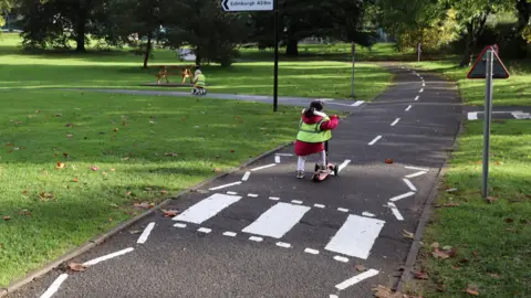 The new cycleway running through Alice Park. The wide path has mini road signs and pedestrian crossings, and children can be seen practicing on the path on small bikes and scooters. 