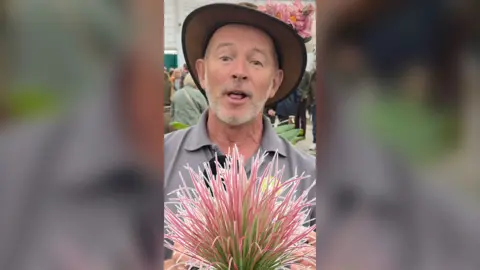 A man with a shirt white beard stood in front of a pink and green plant