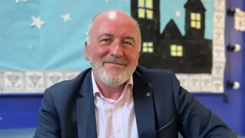 Kurt Roosen, a bald man with a grey beard smiles, he wears a suit, and there is a painting of a house in the background.